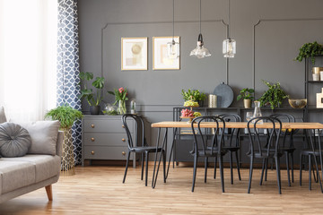 Real photo of a dark flat interior with gray couch, wooden dining table, black chairs and golden posters on the wall in open space living room