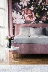 Flowers on white table in pink and grey bedroom interior with pillows on bed. Real photo