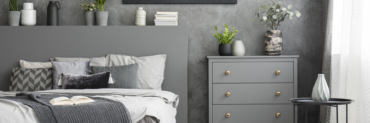 Grey cupboard with fresh plants and ceramic vase standing in simple bedroom interior with books, raw wall and bed with many pillows