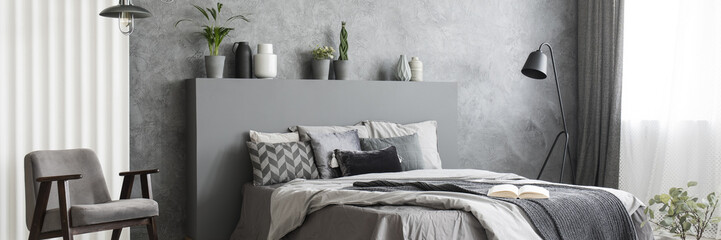 Black metal lamp standing next to double bed with grey sheets in monochromatic bedroom interior with fresh plants, armchair and window with drapes