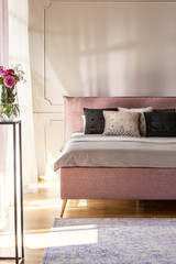 Flowers in pink bedroom interior with bed with cushions against white wall with molding. Real photo