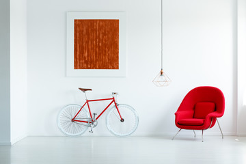 Red bicycle next to armchair in minimal white apartment interior with painting and lamp. Real photo