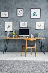 Wooden chair at desk with computer desktop against grey wall with posters in workspace interior....