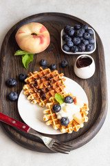 Belgian waffles with fresh blueberries, peaches and chocolate sauce on a rustic wooden cutting board. Liege style belgian waffles. Breakfast or lunch concept.