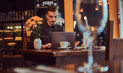 Handsome freelancer man with stylish beard and hair dressed in a black suit working on laptop while sitting at a cafe.