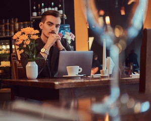 Portrait of a pensive handsome businessman with stylish beard and hair dressed in a black suit sitting with hand on chin at a cafe with an open laptop, lookong at the camera.