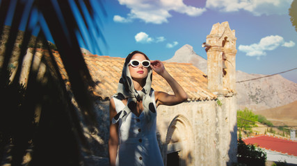 Young girl dressing in 60s style clothes in a village with old church on background. Crete, Greece
