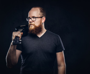 Redhead bearded male musician wearing glasses dressed in a gray t-shirt holds ukulele. Isolated on a dark textured background.