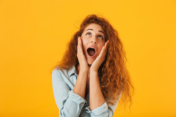 Photo of excited happy woman 20s grabbing face and shouting while looking upward at copyspace, isolated over yellow background
