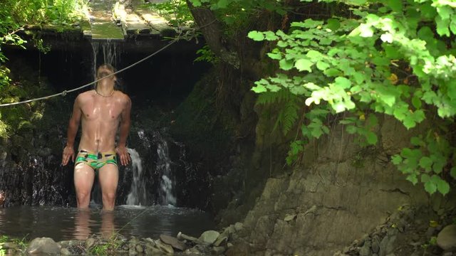 The guy is standing under a small waterfall. EMC on head flows down water. Waterfall in the forest. On the background of trees and forests.