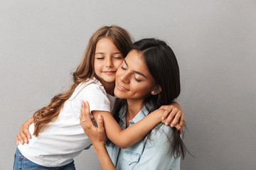 Photo of cheerful woman with little daughter smiling and hugging together, isolated over gray...