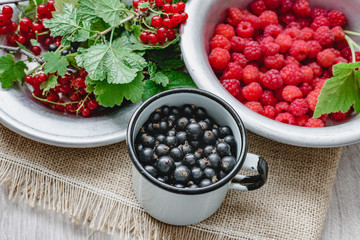Rustic still life of ripe red and black currants, raspberries with leaves in a rustic plate and mug on the background of sacks, healthy food and vitamins. Mixed summer berries.