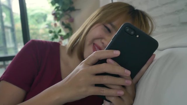 Asian woman playing smartphone while lying on home sofa in her living room. Happy female use phone for texting, reading, messaging and buying online at home. Lifestyle woman at home concept.