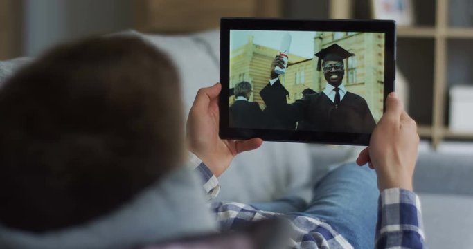 Rear of brunette man lying on the sofa and watching a video of web cam chat on the tablet with an African American male student who just got his diploma on graduation day. Close up.