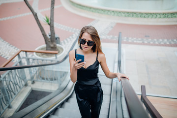 Beautiful young woman with phone hand on an escalator in modern Shopping Mall- music, happiness, technology concept.