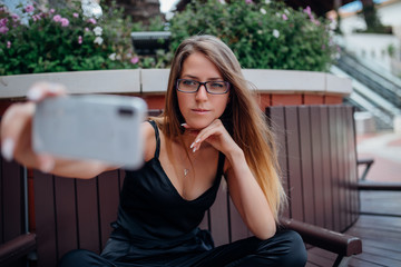 Portrait of a happy young girl while sitting on a bench and taking a selfie outdoors in the city