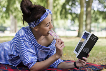 Young woman reading book lying on the grass and eating ice cream