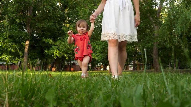 Baby and mom are taking their first steps holding their hands over lawn grass in summer city park and smiling.