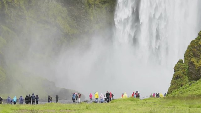 Skogafoss Skogar waterfall bottom, Iceland with white water falling off cliff in green mossy summer, rocky landscape, people walking far in distance on trail with ponchos, umbrellas