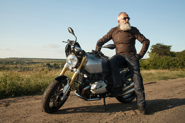 Old rider biker man in black leather jacket, jeans, boots and helmet sit on classic style cafe...