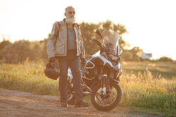 Fototapeta premium Old bearded Biker standing in front of a motorcycle and looking away outdoors