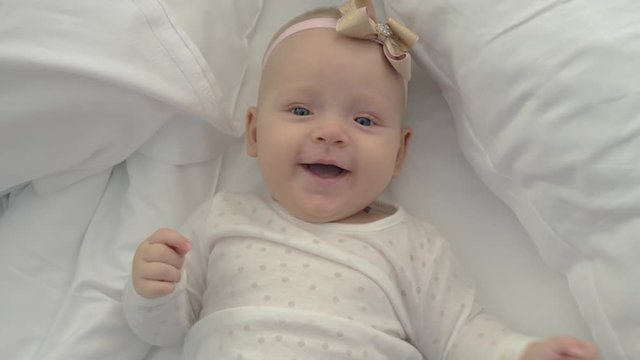 An adorable baby girl is lying on a bed on white sheets. She is laughing happily. A pink and golden bow on her head and a polka dots blouse are making her even cuter