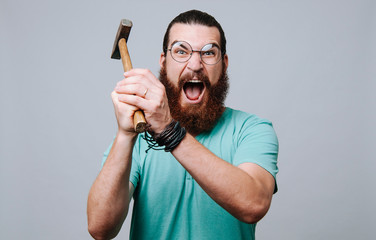 Bearded young man looking and screaming at the camera holding a hammer and ready to hit. - obrazy, fototapety, plakaty