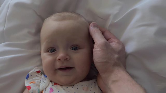 A slow motion of a man's hand gently touching baby girl's head. Six months old girl is lying on her back on light sheets. She is smiling softly feeling father's touch