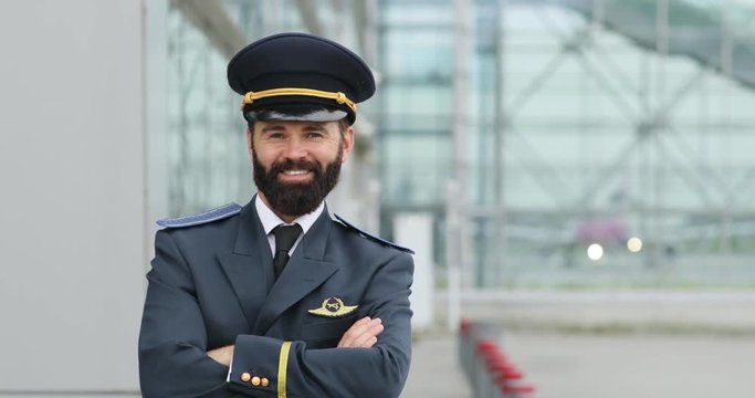 Handsome hot cheerful bearded pilot in the uniform crosses arms on his chest and brightly smiles straight to camera. Confidence, positive emotions, good mood. Joy of life, favorite job. Male portrait