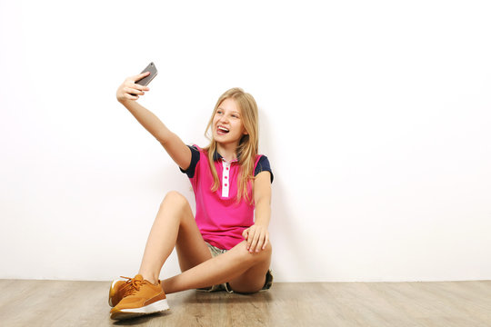 Beautiful teenage girl sitting on wooden floor taking selfie shots on her cell phone. Casual young female in yellow sneakers photographs herself on smartphone, leaning on wall. Background, copy space.