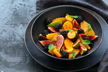 Fruit salad with fresh blackberries, figs and kumquats on a plate on black background. Healthy eating.