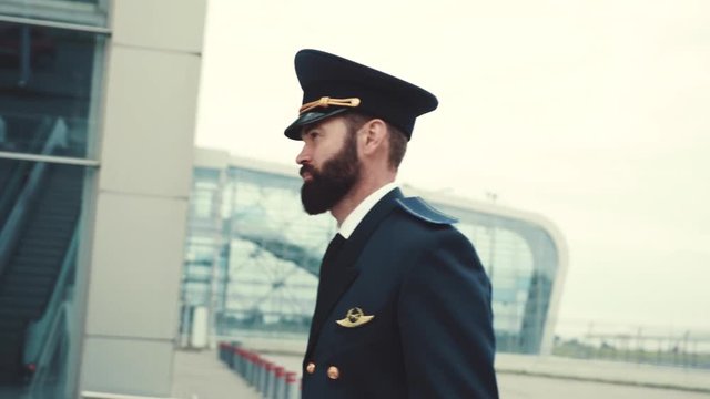 Handsome bearded pilot in formal neat uniform puts on cap, and pulls a suitcase to the plane. Going to work, profession concept, hurrying up. Active lifestyle, luxury. Male portrait