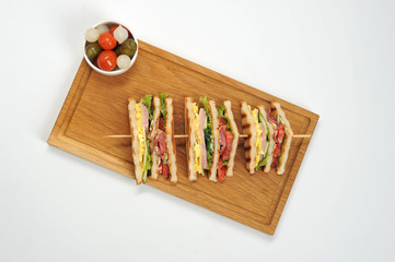 Sandwich on a wooden tray. The sandwich is cut into four pieces and strung on a wooden skewer. Next to a plate of pickles-marinated onions, tomato, cucumbers. White background. View from above.