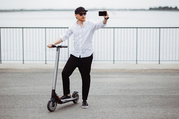 Modern man in stylish outfit making selfie while standing at the street with electric scooter