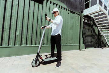 Modern man in stylish outfit using his smartphone while standing at street with electric scooter