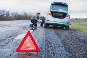 The man is changing the damaged wheel of the car on the roadside of a bad road after the rain against the background of an orange  emergency  triangle warning