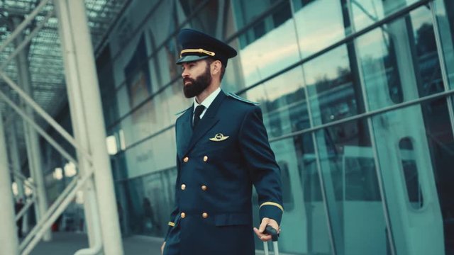 Handsome caucasian bearded man in pilot uniform stands by the airport terminal, puts on the cap, pulls his luggage and leaves the terminal. Profession concept. Male portrait