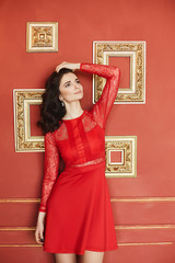 Fashionable and sexy brunette model girl with curly hair in a red stylish dress adjusting her hairstyle and posing at luxury vintage interior