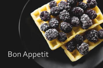 Belgian waffles with frozen blackberries on a black background, inscription bon appetit.
