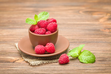 Fresh ripe raspberries with green mint leaves in brown cup and saucer on sackcloth and wooden background.