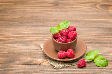 Fresh ripe raspberries with green mint leaves in brown cup and saucer on sackcloth - close up macro photography.