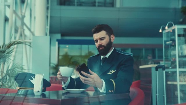 Lonely handsome pilot sits in caf&eacute;, drinks coffee, receives the message, checks his phone and cheerfully laughing at it. Profession concept, lunch, break. Waiting for departure. Male portrait
