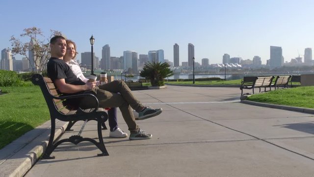 Couple relaxing on city bench in front of the skyline at morning time. In the background is the downtown San Diego with skyscrapers. Concept about love, relationship, and travel.