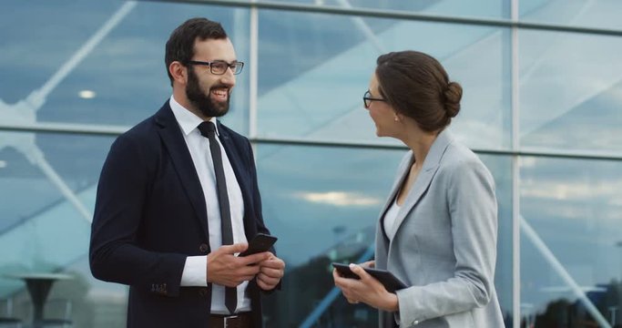 Good looking man in the business style and smartphone in hands speaking and making compliments to the pretty woman in official style. Outdoor their office.