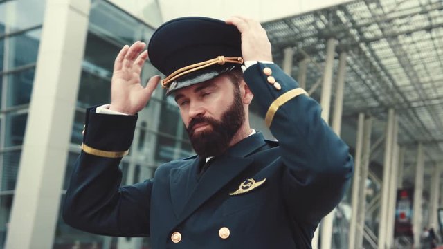 Close up stylish handsome bearded pilot puts on his pilot cap, corrects the uniform and confidently leaves the airport. Day off, weekend, profession concept. Male portrait