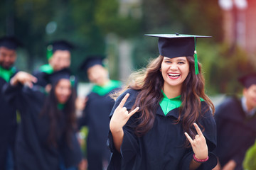 portrait of smiling successful indian student in graduation gown with rock n roll hand gesture