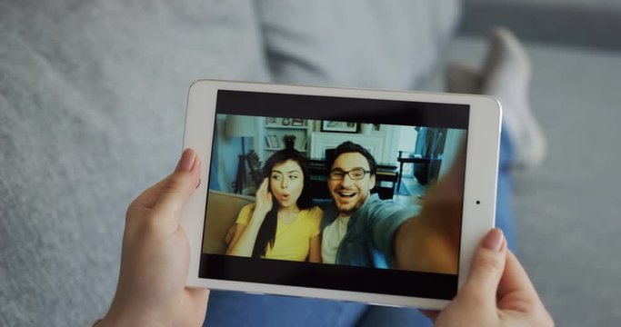 View Over The Shoulder On The Caucasian Couple On The Tablet Screen Videochatting With A Caucasian Woman Who Lying On The Couch At Home. Rear. Close Up.