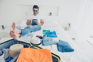 Time of holidays! Full-legh full-body portrait of happy young man looks at the tablet laughs and buys tickets for a trip to the ocean