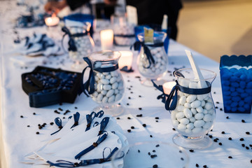 Tavola con confetti, bicchieri, brocche, candele e nastri blu per una cerimonia italiana, all'ombra...