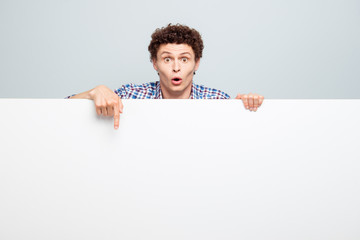 Cheerful young man standing behind the white blank banner and pointing down at a white copyspace isolated on light gray background